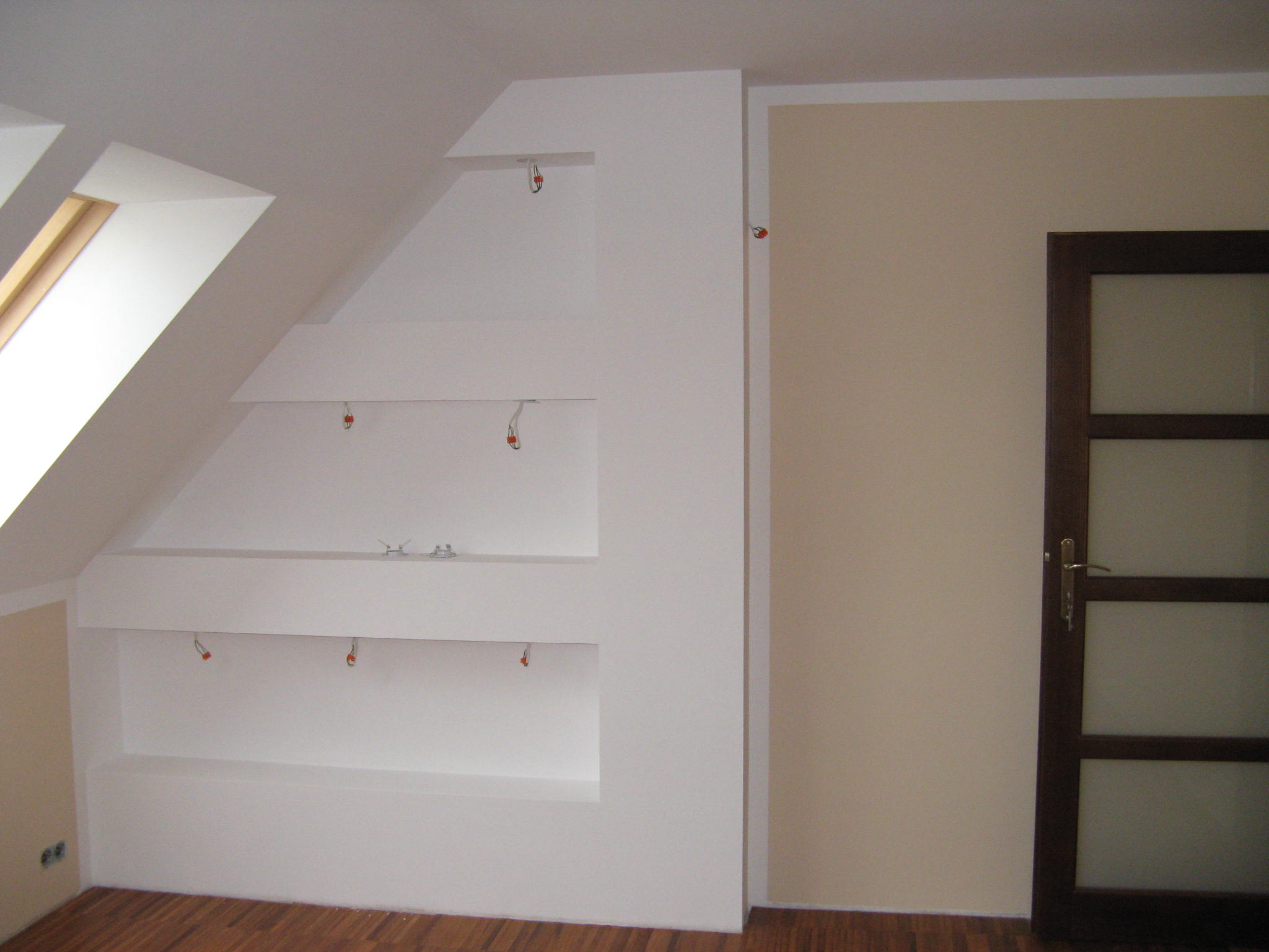 Biała zabudowa wnęk na poddaszu z przygotowanymi kablami do oświetlenia, widoczne okno dachowe i drewniana podłoga.